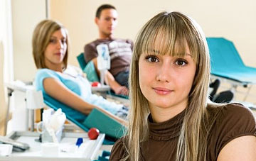 Foto: Eine junge Frau schaut in die Kamera - im Hintergrund zwei Personen bei der Blutspende.